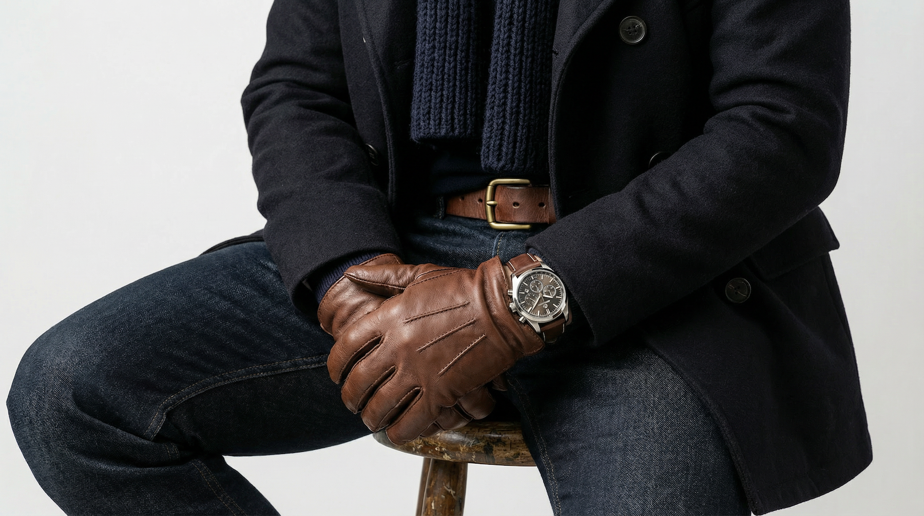 Middle-aged man in navy overcoat, knitted scarf and brown leather gloves sitting on a stool – men's accessories styling for GRAND London